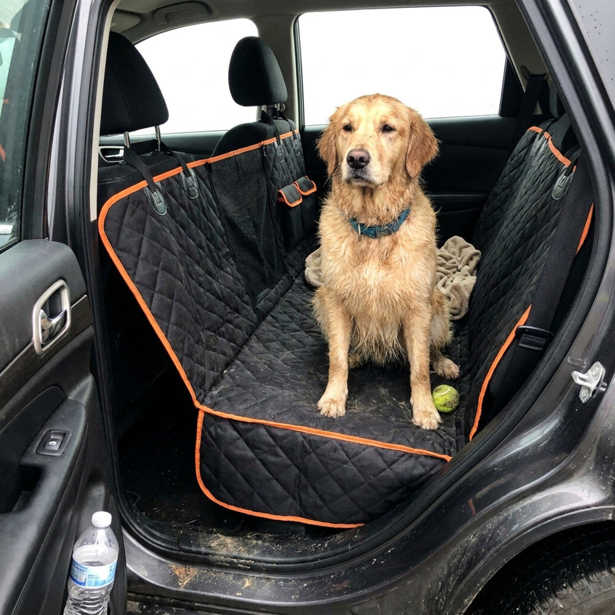 A Golden Retriever sits on a black quilted hammock seat cover with orange trim in the back of an SUV, a tennis ball and a water bottle nearby, photographed from the open rear door