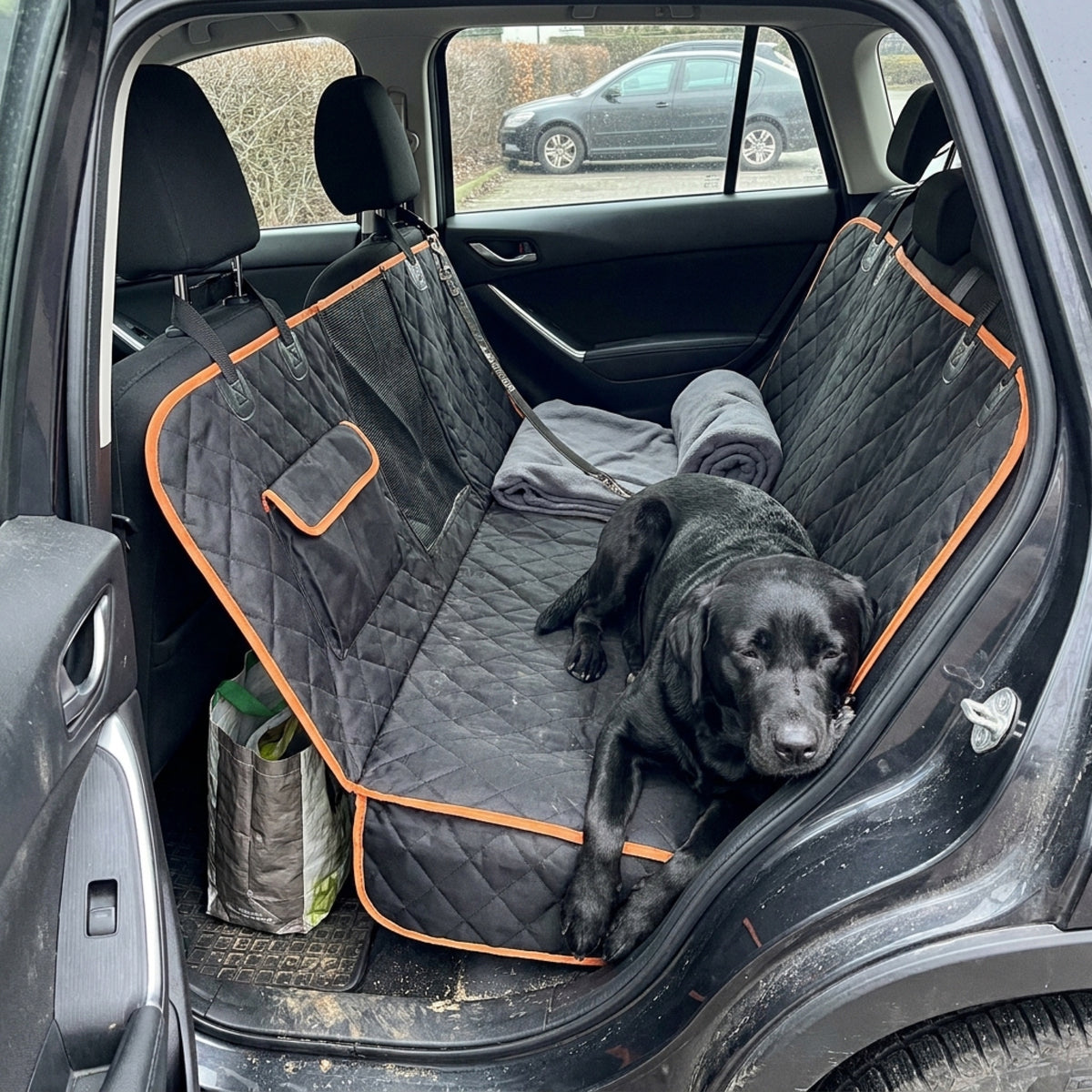 A black Labrador lies across a black quilted hammock seat cover with orange trim, chin on the raised edge, a rolled blanket and a shopping bag beside it