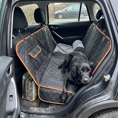 A black Labrador lies across a black quilted hammock seat cover with orange trim, chin on the raised edge, a rolled blanket and a shopping bag beside it